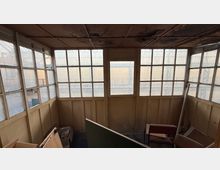 Innenraum einer verwitterten, verglasten Hütte oder eines Warteraums mit großen, in Raster unterteilten Fenstern an drei Seiten und einer Tür in der Mitte. Die Decke ist fleckig und beschädigt, im Raum stehen verstreute Möbelteile und Bretter.