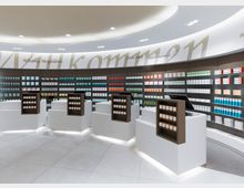 Innenraum einer modernen Apotheke mit geschwungener Regalwand, auf der zahlreiche bunte Medikamentenverpackungen angeordnet sind. Im Vordergrund stehen weiße Verkaufstheken mit eingebauten Regalen für Produkte, beleuchtet durch eine helle Deckenbeleuchtung.