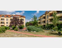 Zwei gelb verputzte Wohngebäude mit roten Ziegeldächern umrahmen einen begrünten Innenhof, der mit einem kleinen Gehweg, Sitzbänken und Bäumen gestaltet ist. Im Vordergrund sind Gräser und Sträucher zu sehen, während der Himmel klar und leicht bewölkt ist.