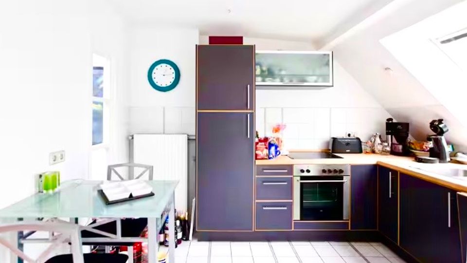 Helle Dachgeschossküche mit schrägen Decken, weißen Fliesen und L-förmiger Einbauküche in dunklen Fronten mit Holzarbeitsplatte, Herd und Spüle. Links steht ein kleiner Glastisch mit Stühlen, dazu sind Fenster und Hängeschränke an den Wänden zu sehen.