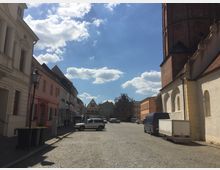 Eine gepflasterte Straße in einer historischen Altstadt, gesäumt von Häusern mit verputzten Fassaden, teils in Pastellfarben. Rechts ist ein großer Kirchturm aus rotem Backstein sichtbar; im Hintergrund stehen renovierte Gebäude und ein Gerüst.