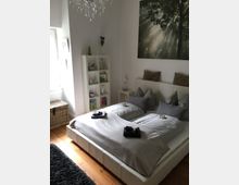 Schlafzimmer mit einem Doppelbett mit grauer Bettwäsche, mehreren Kissen und kleinen Handtüchern auf der Bettdecke. Im Hintergrund befindet sich ein Regal mit Deko-Elementen, eine Lampe, ein Bild von Waldlichtstrahlen und ein Fenstersims mit natürlichem Licht.