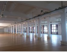 Weitläufiger, leerer Innenraum eines Gebäudes mit einer Reihe weißer Säulen, einem Boden aus hellem Holz und großen Fenstern mit roten Vorhängen. Die Decke ist hoch und mit sichtbaren Schienen und Beleuchtung ausgestattet.