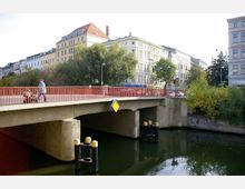 Straßenbrücke mit roten Geländern über einem Fluss in einer städtischen Umgebung, umgeben von mehrstöckigen Wohn- und Geschäftshäusern sowie Bäumen. Im Vordergrund sind Spundpfähle im Wasser sichtbar, während auf der Brücke eine Person mit Kinderwagen entlanggeht.