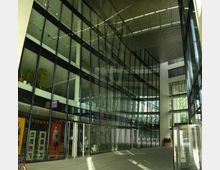 Moderne Glasfassade eines Gebäudes mit einem überdachten Innenhof. Die Wände bestehen hauptsächlich aus großen Fenstern, durch die Tageslicht einfällt, und auf der unteren Ebene befinden sich Geschäfte mit gläsernen Eingängen.