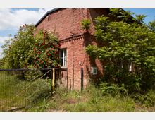 Ziegelhaus mit verwitterter Fassade, umgeben von dichtem Grün und blühenden Rosensträuchern. Ein Drahtzaun mit offener Tür grenzt den bewachsenen Vorgarten ab, während das Haus Fenster mit weißen Rahmen und teils sichtbare Stromanschlüsse aufweist.