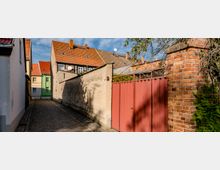 Schmale gepflasterte Gasse mit Häusern, deren Fassaden beige, grün und weiß gestrichen sind, sowie roten Ziegeldächern. Rechts im Vordergrund eine hohe Backsteinmauer mit rotbraunem Holztor, umgeben von Pflanzen.