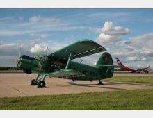 Grünes Doppeldecker-Flugzeug auf einem Flugfeld mit Betonfläche, im Hintergrund grasbewachsene Flächen und ein Verkehrsflugzeug auf einer Start- oder Landebahn, bei leicht bewölktem Himmel.