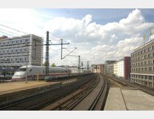 Ein moderner ICE-Zug fährt auf erhöhten Gleisen durch eine Stadtlandschaft. Umgeben von mehrstöckigen Gebäuden stehen Strommasten und Oberleitungen entlang der Schienen, unter einem Himmel mit vereinzelten Wolken.