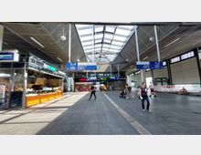 Heller, moderner S-Bahnhof mit verglastem Dach, das viel Tageslicht hereinlässt. Zu sehen sind Hinweisschilder zu den Gleisen, ein kleiner Imbissstand sowie Menschen, die sich durch den Bereich bewegen.