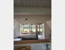 Blick aus einem Wohnraum mit sichtbaren weißen Deckenbalken auf ein großes Fenster und eine geöffnete Tür zur Terrasse. Draußen sind eine überdachte Sitzgruppe, Steinplatten und ein grüner Garten mit Holzzaun zu sehen.