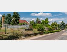 Ein ländlicher Straßenzug mit einem Gewächshaus und einem Garten auf der linken Seite, umgeben von Bäumen, Sträuchern und einem Maschendrahtzaun, unter klarem, blauem Himmel. Im Hintergrund sind ein Haus mit rotem Ziegeldach und weitere Vegetation zu sehen.