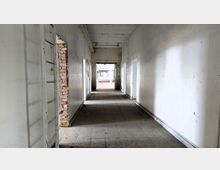 Langer, leerer Flur in einem heruntergekommenen Gebäude mit weißen Wänden, abbröckelndem Putz und sichtbaren Ziegelsteinen an einer Stelle; der Boden ist gefliest und Tageslicht fällt durch Türen und Fenster im Hintergrund.