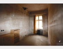 Eine kleine, unrenovierte Wohnung mit nackten, abgeschabten Wänden und einem fleckigen Boden. Ein Fenster mit gelblichem Vorhang lässt etwas Licht herein, daneben steht ein kleiner Heizkörper; links befinden sich zwei einfache Holzregale.