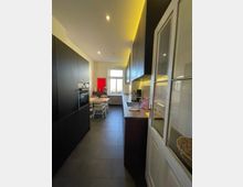 Moderne Küche mit dunklen Schränken, Holzarbeitsplatte und integrierter Beleuchtung. Im Hintergrund ein Esstisch mit Stühlen vor einem Fenster mit weißem Rahmen; der Boden besteht aus dunklen Fliesen.