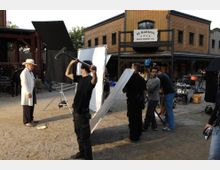 Eine Filmszene auf einem Set, das eine Westernstadt darstellt; im Hintergrund eine Holzkulisse mit einem Gebäude, auf dem ein Schild mit der Aufschrift "US Marshal" zu sehen ist. Im Vordergrund arbeiten mehrere Crewmitglieder mit Beleuchtung, Mikrofonen und Equipments, während eine Person in westernähnlicher Kleidung steht.