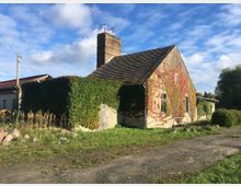Ein altes Haus mit grauem Satteldach und einem hohen Schornstein, teilweise von grünen und roten Kletterpflanzen bedeckt. Es steht in einer ländlichen Umgebung mit einem Feldweg im Vordergrund und Bäumen im Hintergrund.