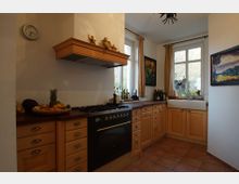 Eine helle, traditionelle Küche mit Holzschränken, einem schwarzen Backofen mit Gasherd und einem großen Spülbecken unter einem zweiflügeligen Fenster. Auf der Arbeitsfläche stehen Obstkorb, Gewürze und Flaschen, die Wände sind mit Gemälden dekoriert.