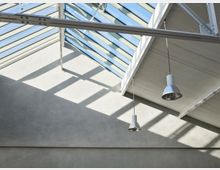 Ein Raum mit einer schrägen Decke, die teilweise aus Glas besteht und Tageslicht hereinlässt. Zwei industrielle Pendelleuchten hängen von der Decke, und die Wände sind aus glattem, grauem Beton.