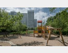 Spielplatz mit hölzernem Klettergerüst und Rutsche auf einem sandigen Untergrund, umgeben von Bäumen und Grünflächen im Vordergrund; im Hintergrund ein mehrstöckiges Wohngebäude.