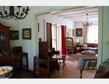 Wohnzimmer mit klassischer Einrichtung, einschließlich Ledersofas, Holzmöbeln und Gemälden an den Wänden. Der Raum ist durch große Fenster mit Vorhängen hell beleuchtet, und ein Kronleuchter hängt an der Decke; ein angrenzender Essbereich ist zu sehen.