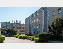 Mehrstöckige Wohngebäude mit schlichten Fassaden, Balkonen und großen Fenstern, umgeben von Büschen und einem gepflasterten Hofbereich. Im Vordergrund befinden sich Grünflächen und ein Schild.