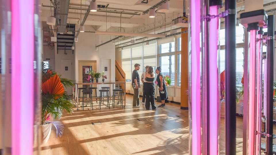 Bright, modern indoor space with wood floors, exposed ceiling pipes, and large windows along one wall letting in sunlight. Tall vertical pink LED grow lights and potted plants frame a high table with stools, giving the room an urban greenhouse or studio feel.