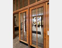 Holztür mit großen Glaseinsätzen, die zu einem hellen Empfangsbereich mit Marmorboden, hohen Säulen und einer geschwungenen Treppe führt. Im Hintergrund sind Sitzmöbel, Pflanzen und ein Deckenleuchter sichtbar.