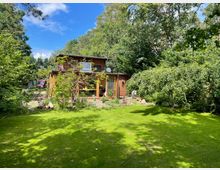 Holzhaus mit großen Fenstern und Flachdach, umgeben von einem üppig bepflanzten Garten mit Rasen, Bäumen und Sträuchern. Im Hintergrund dichter Wald und blauer Himmel.