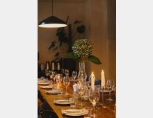 Langer, gedeckter Holztisch mit Tellern, Gläsern und Besteck, dekoriert mit einer Vase mit Hortensien und brennenden Kerzen, in einem warm beleuchteten Innenraum mit Pflanzen im Hintergrund.