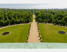 Luftaufnahme eines symmetrischen Gartens mit breitem Sandweg in der Mitte, flankiert von Bäumen und dekorativen Pflanzen. Die großen, gepflegten Rasenflächen enthalten runde Brunnen, und im Hintergrund erstreckt sich ein dichter Wald bis zur Skyline einer Stadt.
