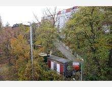 Blick auf einen kleinen Schrebergarten mit einem Holzhaus und umliegendem Herbstlaub, angrenzend an einen gepflasterten Weg. Im Hintergrund sind mehrstöckige Wohnhäuser mit hellen Fassaden sichtbar.