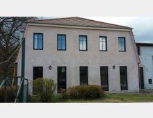 Zweistöckiges Gebäude mit hellem Putz, schwarzen Fenstern und rotem Ziegeldach. Vor dem Haus befinden sich niedrige Büsche und eine Grünfläche, daneben ist ein Teil eines angrenzenden Gebäudes sichtbar.