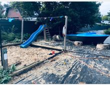 Spielplatz mit einem Sandkasten und einer blauen Rutsche, daneben steht ein kleines blaues Boot auf einem mit schwarzem Vlies bedeckten Boden. Im Hintergrund Bäume, ein Gartenzaun und Häuser.