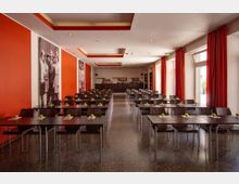 Großer Veranstaltungsraum mit dunklen Holzstühlen und Tischen, die in Reihen aufgestellt sind, jeweils dekoriert mit einem grünen Apfel und Papierblättern. Die Wände sind in Orange und Weiß gestaltet, mit großen Schwarz-Weiß-Fotografien, während bodentiefe Fenster mit roten Vorhängen Tageslicht hineinlassen.