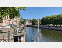 Eine Uferpromenade entlang eines ruhigen Kanals mit einer steinernen Brücke im Hintergrund. Links steht ein historisches Gebäude mit rosa Fassade, während rechts Bäume eine Allee bilden.