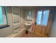 Ein einfaches Badezimmer mit weiß gefliesten Wänden, einem Wandwaschbecken mit Spiegel darüber, einem Handtuchhalter-Heizkörper, einer Holztür mit Glas und Vorhängen sowie einem kleinen Fenster mit Fensterbank.