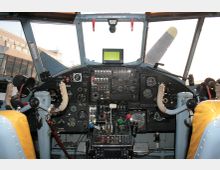 Cockpit eines älteren Flugzeugs mit umfangreichen analogen Instrumenten, Schaltern und Hebeln. Durch die Frontscheibe sind ein Propellerblatt und ein Flughafengebäude im Hintergrund erkennbar.