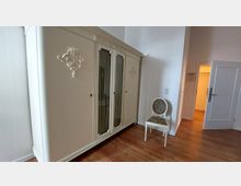 Großes, cremefarbenes Vintage-Schranksystem mit floralen Verzierungen, daneben ein weißer Stuhl mit gepolsterter Sitzfläche im floralen Design; das Zimmer hat Holzboden und helle Wände, im Hintergrund eine offenstehende Tür zu einem weiteren Raum.