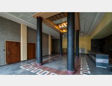 Innenansicht eines Eingangsbereichs mit schwarzen Säulen, einem gemusterten Marmorboden und einer repräsentativen Deckenleuchte. An den Wänden befinden sich zwei Holztüren, gestrichene Wände sowie ein Tresen mit schwarzer und roter Marmorverkleidung.