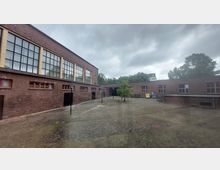 Eingezäunter Hof mit Gebäuden aus rotem Backstein, großen modernen Fenstern und einer zentralen Grünfläche mit einem einzelnen Baum. Der Himmel ist bewölkt, und im Hintergrund sind Bäume sowie Wohnhäuser zu erkennen.