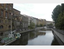 Kanalansicht in einer Stadt mit Backsteingebäuden links und modernen Wohnhäusern rechts; ein Boot ist am Ufer vertäut, und im Hintergrund führt eine Brücke über den Kanal. Entlang des Wassers verlaufen Gehwege, gesäumt von Bäumen und Geländern.
