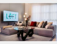 Modernes Wohnzimmer mit einer großen grauen Eckcouch, dekoriert mit bunten Kissen. Im Vordergrund steht ein schwarzer Couchtisch mit geschwungener Basis, daneben eine Stehlampe mit mehreren Kugelleuchten, und im Hintergrund ein Fernseher mit der Aufschrift „LÖFT“.