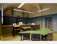 Innenansicht eines historischen Gerichtssaals mit hoher Gewölbedecke. Der Raum ist mit dunkelgrünen Holzvertäfelungen, verzierten Wandmalereien, einer Holzrichterbank und einem grünen Besprechungstisch mit grauen Stühlen ausgestattet.