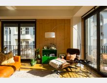 Wohnzimmer mit großen bodentiefen Fenstern, die viel Tageslicht hereinlassen, Holzwandverkleidung, einem orangefarbenen Ledersofa, einem braunen Lounge-Sessel mit Fußhocker, einer Stehlampe und einem grünen Sideboard. Im Hintergrund sind Balkone eines gegenüberliegenden Gebäudes sichtbar.