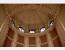 Innenansicht einer Kuppel in einem historischen Gebäude mit sternförmigen Verzierungen auf der gewölbten Decke. Rundfenster mit farbigen Glasmalereien und Säulenelementen rahmen die Struktur ein, während die Wände in warmen Farben mit detaillierten Ornamenten gestaltet sind.