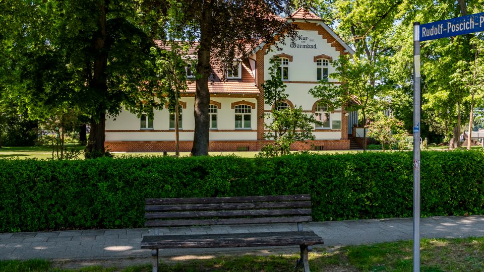 Blick auf ein historisches Kurgebäude mit weißer Fassade und rotem Ziegeldekor, umgeben von hohen Bäumen und Grünanlagen. Im Vordergrund stehen eine Parkbank, eine Hecke entlang des Gehwegs und ein Straßenschild „Rudolf-Poschich-Platz“.