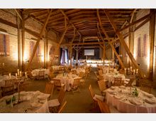 Innenraum einer rustikalen Veranstaltungslocation in einer Scheune mit freiliegenden Backsteinwänden und Holzbalken. Der Raum ist für ein Fest eingedeckt, mit runden, weiß gedeckten Tischen, Holzkohlefackeln und Kerzenleuchtern.