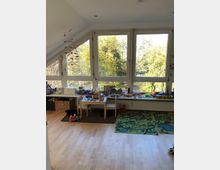 Ein helles Kinderzimmer mit großen Fenstern, durch die Bäume und Tageslicht sichtbar sind. Im Vordergrund sind bunte Spielmatten, Spielzeugkisten, ein kleiner Tisch mit Stühlen sowie eine Fensterbank mit Spielzeug und Bastelmaterialien zu sehen.