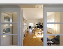 Wohnzimmer mit Holzparkettboden, einem langen Holzesstisch und schwarzen Stühlen. Im Hintergrund befinden sich ein schwarzer Flügel, eine große Bücherwand und helle Fenster mit lichtdurchfluteten Vorhängen.